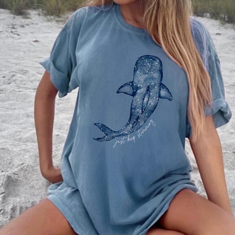 Puede incluir: Camiseta azul con una ilustraci&oacute;n de ballena. La ballena es de color azul oscuro con motas blancas, y el texto "just keep swimming" est&aacute; escrito en cursiva debajo.