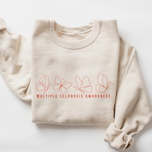May include: A cream-colored sweatshirt with a red line drawing of four butterflies. The words "MULTIPLE SCLEROSIS AWARENESS" are printed in red below the butterflies. The sleeves are folded.