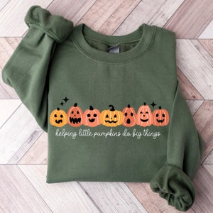 May include: A green sweatshirt with a line of orange and brown jack-o'-lantern faces and the text "helping little pumpkins do big things".