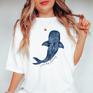 Puede incluir: Camiseta blanca con un dise&ntilde;o de ballena azul y un patr&oacute;n de estrellas. La ilustraci&oacute;n de la ballena tiene las palabras "just keep swimming" en una fuente manuscrita. La camiseta es de estilo casual y c&oacute;modo.