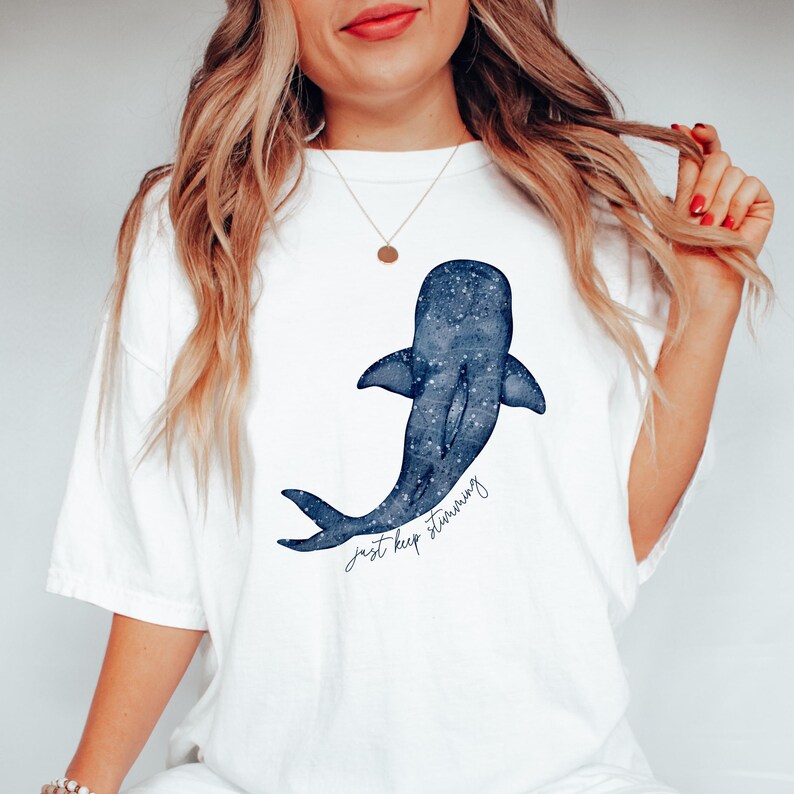Puede incluir: Camiseta blanca con un dise&ntilde;o de ballena azul y un patr&oacute;n de estrellas. La ilustraci&oacute;n de la ballena tiene las palabras "just keep swimming" en una fuente manuscrita. La camiseta es de estilo casual y c&oacute;modo.
