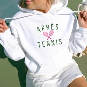 Może przedstawiać: Biała bluza z kapturem z zielonym nadrukiem "APRÈS TENNIS" i różowym wzorem rakiety tenisowej.