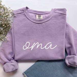 May include: A lavender sweatshirt with the word "oma" written in white cursive on the front.