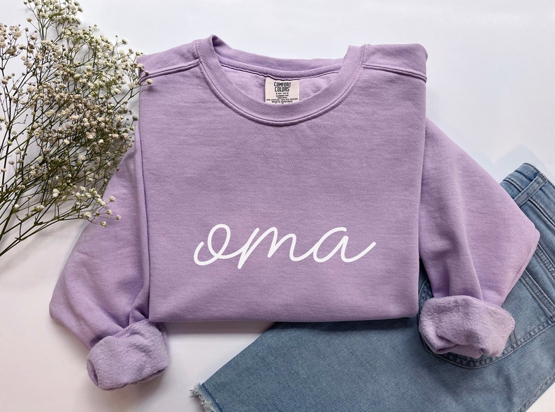 May include: A lavender sweatshirt with the word "oma" written in white cursive on the front.