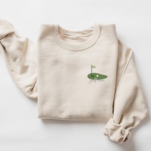 Może przedstawiać: Beżowa bluza z zielonym nadrukiem pola golfowego i napisem "golf mom" z przodu.