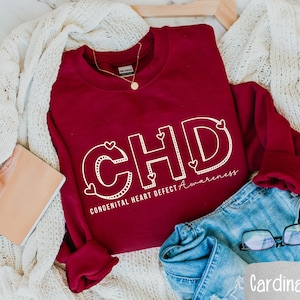 May include: A maroon sweatshirt with the text "CHD CONGENITAL HEART DEFECT AWARENESS" in white outlined letters with small hearts around the letters. The sweatshirt is folded and lying on a white knitted blanket with a pair of blue jeans and glasses.