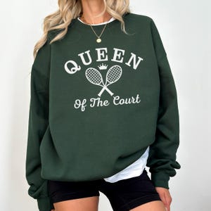 Op de afbeelding: Een donkergroene sweatshirt met de witte tekst "QUEEN of The COURT". Het ontwerp bevat een kroon en gekruiste tennisrackets. De sweatshirt heeft lange mouwen en een ronde hals.