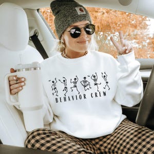 Puede incluir: Sudadera blanca con un gráfico negro de esqueletos bailando y el texto "Behavior Crew".