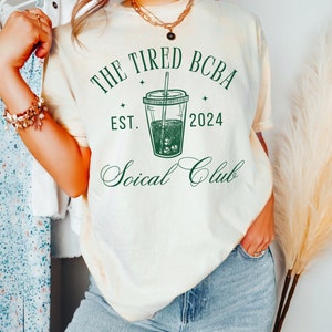 May include: A white t-shirt with a green graphic design that says "The Tired BCBA Est. 2024 Soical Club". The graphic features a stylised drawing of a cup of iced coffee with a straw.