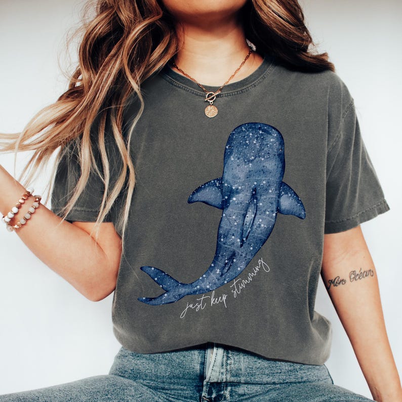 Puede incluir: Camiseta gris oscuro con un dise&ntilde;o de ballena azul y un patr&oacute;n de estrellas. La ilustraci&oacute;n de la ballena tiene las palabras "just keep swimming" en una fuente manuscrita. La camiseta es de un estilo c&oacute;modo e informal.