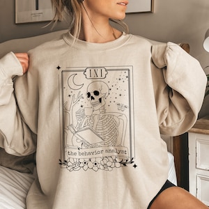 Peut inclure: Un sweat-shirt beige avec une illustration noire et blanche d'un squelette tenant une tasse et un livre. L'illustration est dans le style d'une carte de tarot et comprend le texte "IXI the behavior analyst".