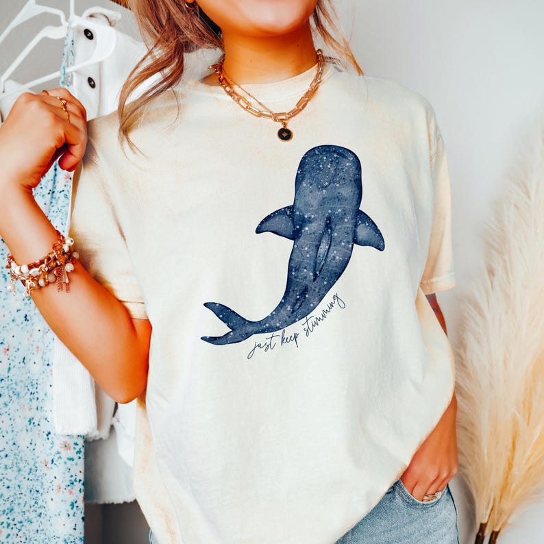 Puede incluir: Camiseta color crema con una ilustraci&oacute;n de ballena azul y un patr&oacute;n de estrellas. El dise&ntilde;o de la ballena va acompa&ntilde;ado del texto manuscrito "just keep swimming". La camiseta tiene mangas cortas y un corte holgado.