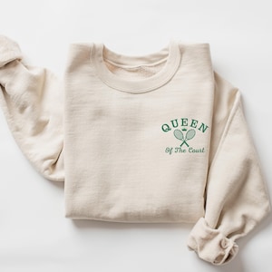 Op de afbeelding: Een crèmekleurige sweatshirt met de tekst "QUEEN Of The Court" in groen, samen met een kroon en gekruiste tennisrackets. De sweatshirt is opgevouwen, met de mouwen zichtbaar.