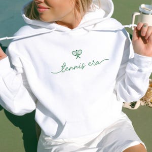 Puede incluir: Sudadera con capucha blanca con bolsillo delantero y cordón. Presenta un gráfico de raqueta de tenis verde y la frase "tennis era" en cursiva. Forma parte de una colección de ropa con temática de tenis.
