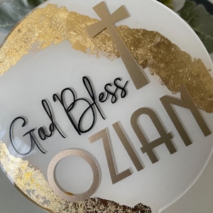May include: White round cake topper with gold leaf accents. The topper features a gold cross, the words "God Bless" and the name "Ozian".
