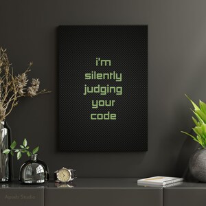 May include: A black framed print with the words "I'm silently judging your code" in green. The artwork is displayed on a dark grey surface, alongside vases, a watch, and a notebook. The theme is tech and humour.