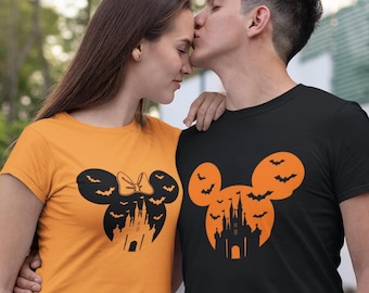Puede incluir: Camisetas a juego para Halloween. Una es naranja, la otra negra. Cada una presenta una silueta de Mickey Mouse con un diseño de castillo y murciélagos. La camiseta naranja tiene un lazo. Ambas camisetas son de manga corta.
