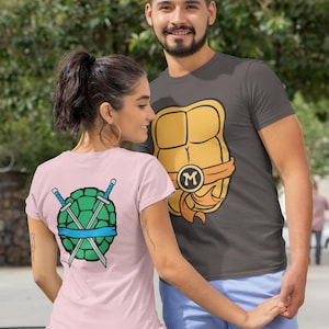 May include: A couple wearing matching t-shirts with a Teenage Mutant Ninja Turtles theme. The man's shirt features a yellow and orange turtle shell with the letter "M" on it. The woman's shirt features a green turtle shell with two crossed swords and a blue bandana.