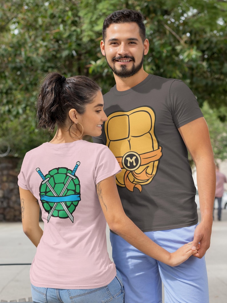 May include: A couple wearing matching t-shirts with a Teenage Mutant Ninja Turtles theme. The man's shirt features a yellow and orange turtle shell with the letter "M" on it. The woman's shirt features a green turtle shell with two crossed swords and a blue bandana.
