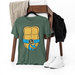 May include: A green t-shirt with a yellow and blue Teenage Mutant Ninja Turtles shell graphic. The shell has a blue bandana with a yellow letter L on it.