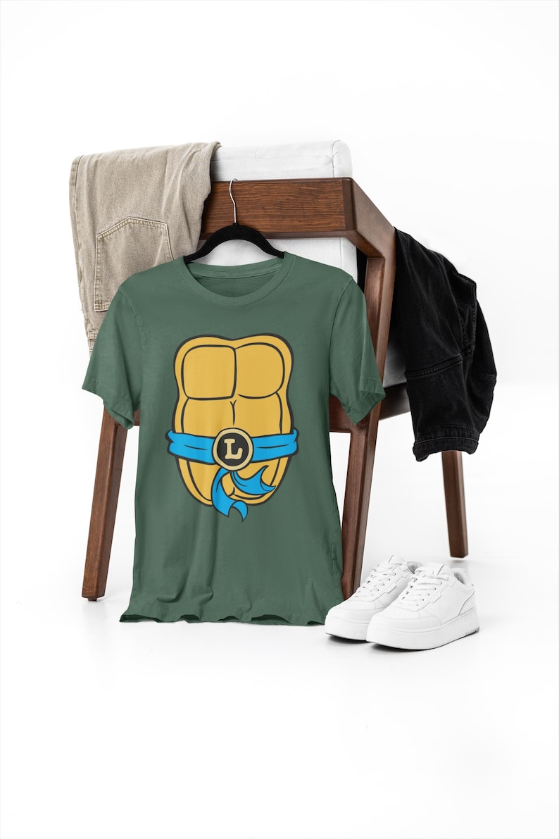 May include: A green t-shirt with a yellow and blue Teenage Mutant Ninja Turtles shell graphic. The shell has a blue bandana with a yellow letter L on it.