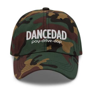 May include: A camouflage baseball cap with the word "DANCEDAD" in white block letters, with "pay-drive-clap" below. The cap features a curved brim and a black button on top. The camouflage pattern includes shades of green, brown, and black.