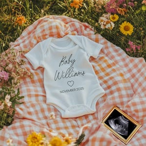 May include: A white onesie with the text "Baby Williams" and a heart, along with the date "November 2025". The onesie is lying on a pink and white checkered blanket surrounded by flowers.