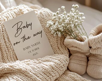 以下が含まれることがあります： 白いカードに黒い書体で「Baby on the way」と書かれており、その下に「Baby Smith October 2025」というテキストがあります。カードはクリーム色のニットブランケットの上に置かれ、白いベビーブレスのブーケとクリーム色のニットのベビーブーティのペアが添えられています。