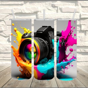 May include: A stainless steel tumbler featuring a vibrant design of a camera lens with colorful paint splashes. The design includes yellow, orange, pink, and blue hues, with a gray background. The tumbler has a silver straw.