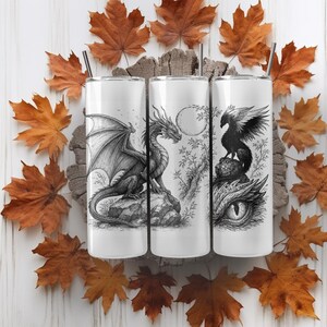 May include: Three white insulated tumblers with black and white fantasy illustrations of a dragon, a dragon with a moon, and a bird. The tumblers have black straws and are surrounded by autumn leaves.