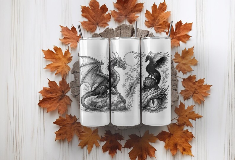 May include: Three white insulated tumblers with black and white fantasy illustrations of a dragon, a dragon with a moon, and a bird. The tumblers have black straws and are surrounded by autumn leaves.