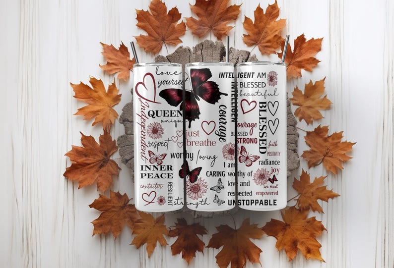 May include: Three white stainless steel tumblers with black straws, decorated with butterflies, hearts, and inspirational words like "Queen", "Blessed", and "Unstoppable". The tumblers are surrounded by autumn leaves.