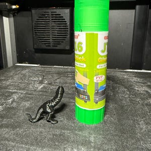 Peut inclure: Une figurine de dinosaure noire imprimée en 3D à côté d'un bâton de colle vert étiqueté "J16 Glue Stick" avec le texte "Pour l'impression 3D, séchage rapide, lavable, lit chaud, PVP NET:30g".
