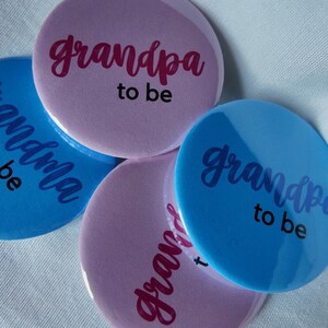 May include: A collection of round buttons in shades of blue and pink. The buttons have the words "grandpa to be" and "grandma to be" in cursive script. The buttons are scattered on a white surface.