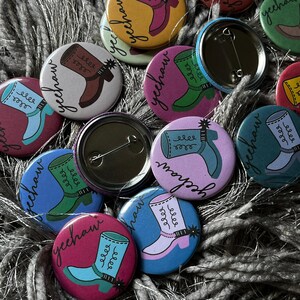 May include: A collection of colorful round buttons with a cowboy boot design and the word "yeehaw" written on them. The buttons are made of metal and have a pin back.