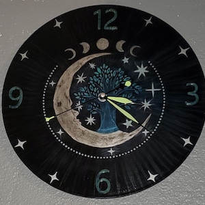 May include: Black round wall clock with a celestial design. The clock face features a crescent moon, a tree, stars, and moon phases. The numbers 3, 6, 9, and 12 are visible. The clock hands are a bright green color.