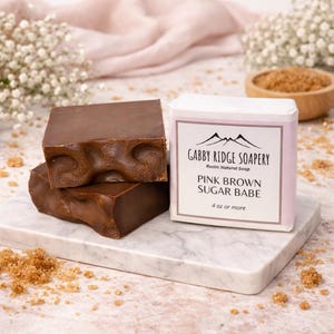 May include: Two brown soaps with a textured surface are stacked on a marble tray. A wrapped soap with the text "Gabby Ridge Soapery" and "Pink Brown Sugar Babe" is next to the soaps. The wrapped soap is 4 oz or more.