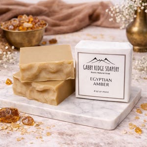 May include: Three bars of light brown soap are stacked on a marble surface. A white box with the text "Gabby Ridge Soapery" and "Egyptian Amber" is next to the soap. The soap is labeled as 4 oz or more.