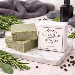 May include: Three green, rectangular soaps with decorative carvings sit on a marble surface, next to a wrapped soap bar. The wrapped bar has the text "Gabby Ridge Soapery" and "The Perfect Man". Rosemary sprigs and peppercorns are scattered around.