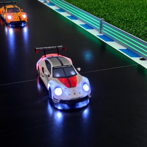 Könnte beinhalten: Zwei ferngesteuerte Porsche-Rennwagen auf einer schwarzen Strecke. Das vordere Auto ist weiß mit einem roten Streifen und leuchtenden Scheinwerfern, das hintere Auto ist orange mit schwarzen Streifen. Beide verfügen über Heckspoiler und Porsche-Schriftzüge.