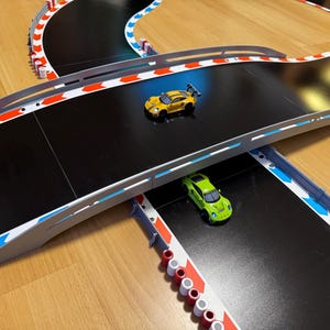 Puede incluir: Una pista de carreras en blanco y negro con bordes rojos y azules. Dos coches de juguete, uno amarillo y otro verde, conducen por la pista. La pista tiene un puente en el medio.