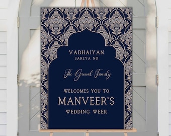 Pode incluir: Uma placa de boas-vindas de casamento azul marinho em um cavalete de madeira. A placa apresenta uma borda floral decorativa e o texto "VADHAIYAN SAREYA NU, The Grewal Family Welcomes You To MANVEER'S WEDDING WEEK."