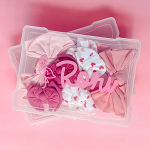 May include: A clear plastic storage container filled with pink baby headbands and bows. A pink flamingo and the name "Rory" are placed inside the container. The background is a solid pink color.