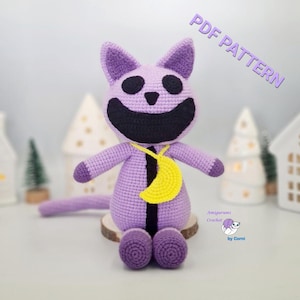 Pode incluir: Boneca de gato de crochê feita à mão em tons de roxo, com um grande sorriso preto e um pingente de lua crescente amarelo. A boneca está sentada em uma fatia de madeira, com o texto "PDF PATTERN" visível. A boneca tem cerca de 20 cm de altura.