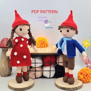Może przedstawiać: Wzór PDF do szydełkowania dziewczęcej i chłopięcej lalki amigurumi gnoma. Lalka dziewczyna ma na sobie czerwoną sukienkę w białe kropki i czerwony kapelusz. Lalka chłopiec ma na sobie niebieski sweter, brązowe spodnie i niebieski szalik. Obie lalki mają brązowe włosy i stoją na drewnianych podstawach.