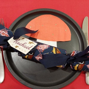 Puede incluir: Un cracker de Navidad azul y blanco con un lazo negro y una etiqueta con el nombre "Rachael". El cracker está en un plato negro con un hongo de papel rojo y blanco.