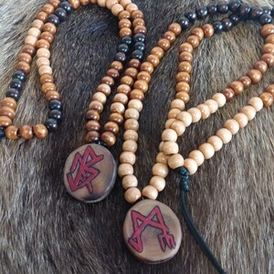 Könnte beinhalten: Halsketten mit Holzperlen und runden Anhängern. Die Anhänger haben eine dunkle Holzoberfläche mit roten Symbolen. Die Perlen sind in Braun- und Schwarztönen gehalten. Die Halsketten werden auf einer strukturierten, braunen und grauen Oberfläche präsentiert.