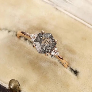 May include: A rose gold engagement ring featuring a hexagonal grey diamond with smaller diamonds set on the band. The ring is displayed in a velvet ring box.