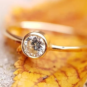 May include: A gold ring with a single round, clear gemstone set in a bezel setting. The ring is resting on a yellow leaf.