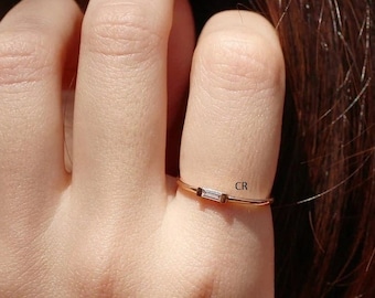 May include: A delicate gold ring with a single, small, rectangular diamond set in the center. The ring is worn on the finger of a person with brown hair.